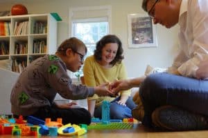 Boy with Down syndrome sits on the floor playing legos with his mom and dad.