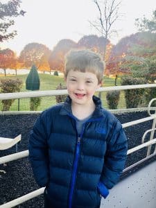 Smiling young boy with Down syndrome standing with hand in pockets of a blue coat.