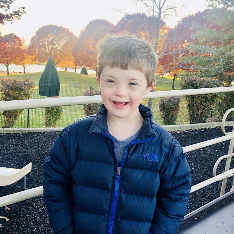 Smiling young boy with Down syndrome standing with hand in pockets of a blue coat.