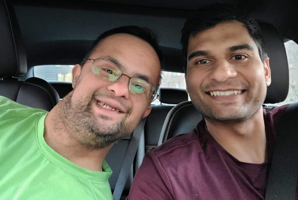 Two men sit side-by-side in a car smiling.